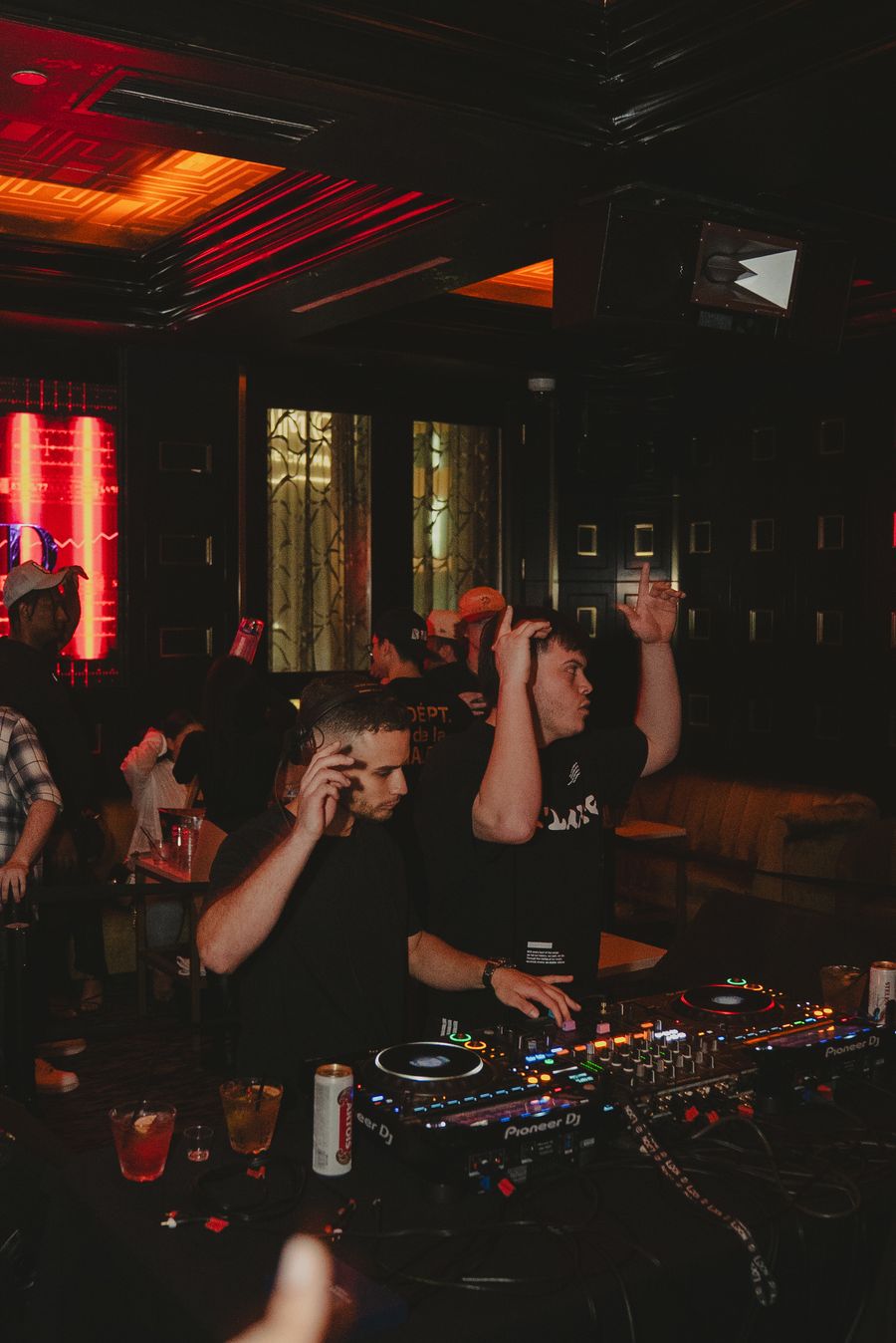 The duo working the mixer under deep red club light, drinks and cables on the booth.